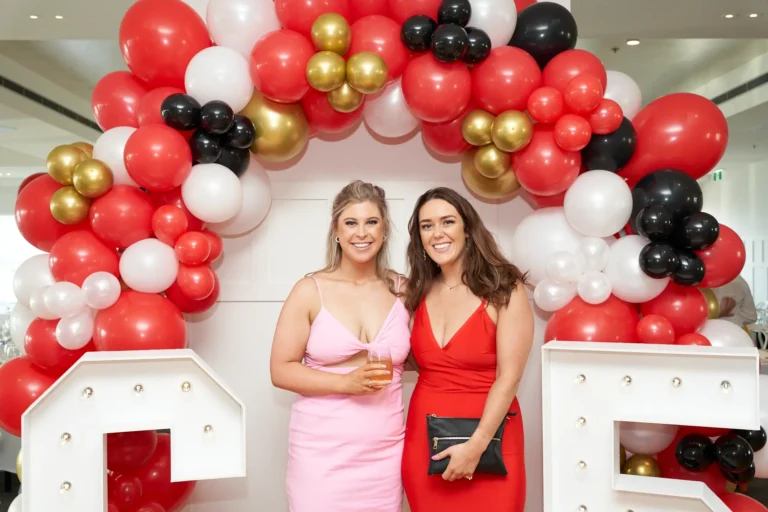 garryowens 65th anniversary ball two women arrival posing Two elegantly dressed women pose together during arrivals at Garryowen’s 65th Anniversary Ball, smiling broadly at the camera. One holds a glass of amber-colored drink, while the other carries a small black clutch. They stand in front of a festive balloon arch in red, white, black, and gold, framed by illuminated letters and numbers as part of the event decor. Polished hardwood floors and softly lit areas with mingling guests complete the modern venue setting. The scene captures a lively, stylish, and celebratory atmosphere, conveying the excitement, glamour, and joyful energy of the awards night reception.