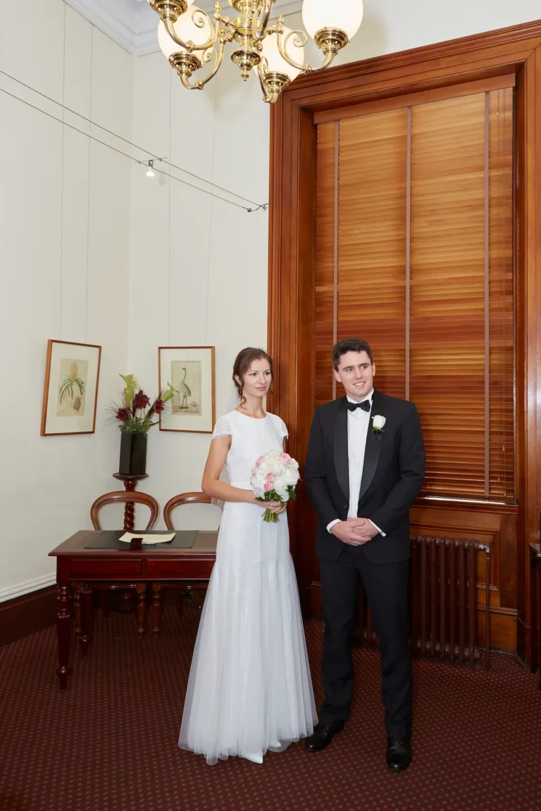 Masha and Marcus stand together in the Thomas Hyde Room of Melbourne’s historic Old Treasury Building, smiling warmly just after completing their civil ceremony. The bride holds her pink-and-white bouquet, while the groom carries their marriage certificate. Behind them, the intimate registry space features dark wood wainscoting, a signing desk with chairs, botanical wall prints, wooden window blinds, and a brass chandelier. The patterned red carpet and soft lighting enhance the room’s heritage charm, capturing a heartfelt and timeless moment moments after they were officially married.