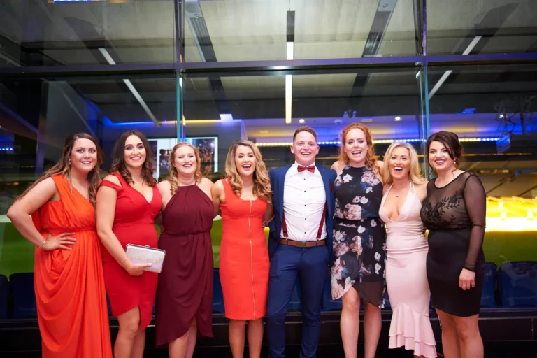 padraig pearses gac awards night mcg elegant group posing stadium window backdrop A group of eight guests—seven women in vibrant evening gowns and one man in a suit with a red bow tie—stand together smiling warmly during the Pearses Awards Night & Gala Dinner at the MCG. Posed confidently before floor-to-ceiling glass windows, they are framed by blue stadium seating and the softly lit green field beyond. The colorful dresses, formal attire, and unique stadium backdrop create a sophisticated, celebratory atmosphere as the group pauses to capture a memorable moment of camaraderie and elegance at the gala.
