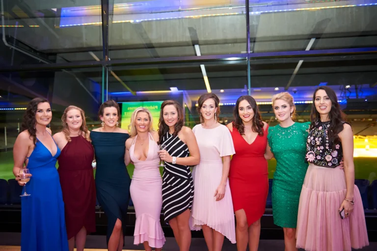 padraig pearses gac awards night mcg elegant women group portrait stadium lighting A group of eight women pose together with warm smiles during the Pearses Awards Night & Gala Dinner at the MCG, dressed in vibrant gowns ranging from blue and maroon to teal, pink, and patterned styles. Their relaxed, confident postures and the wine glass and phone in hand suggest friendly socializing. Behind them, floor-to-ceiling glass reveals dark blue stadium seating and the softly lit green field, enhanced by vivid blue accent lighting. The image conveys an elegant, festive atmosphere, highlighting camaraderie and the unique setting of a gala held inside a major sports arena.