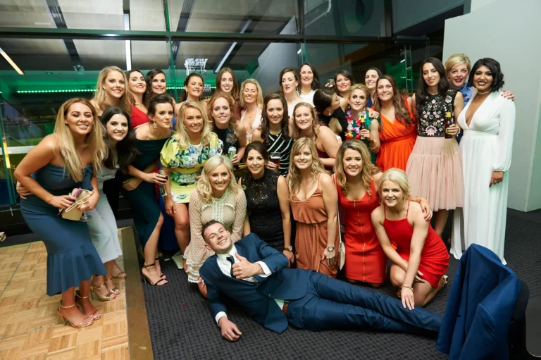 padraig pearses gac awards night mcg group celebration fun A large group of over 20 guests pose energetically during the Pearses Awards Night at the MCG. A man in a blue suit lies on the floor giving a thumbs-up, surrounded by women in vibrant evening gowns, all smiling and holding drinks. Some guests wear leis and raise hands in celebration, capturing a lively, playful, and social atmosphere. The modern function room features floor-to-ceiling glass partitions, dark blue stadium seating beyond, and green ambient lighting reflecting off the glass, emphasizing the festive, high-energy, and memorable nature of the gala event.