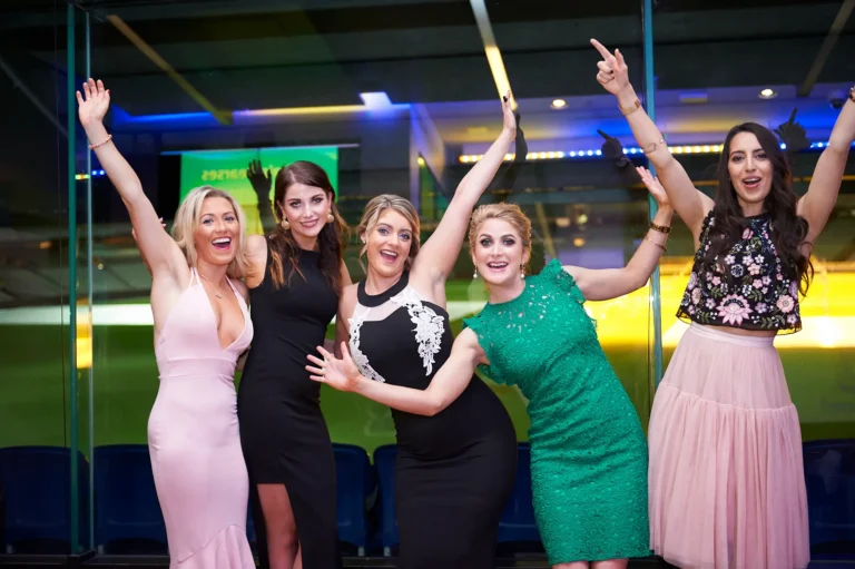 padraig pearses gac awards night mcg joyful women group celebration stadium backdrop A lively group of five women celebrate joyfully during the Pearses Awards Night & Gala Dinner at the MCG, laughing and posing with their hands raised in energetic gestures. Dressed in elegant evening wear, they lean in together, capturing a moment of shared excitement and camaraderie. Behind them, floor-to-ceiling glass reveals dark blue stadium seating and the softly lit green field, enhanced by vibrant blue accent lighting. The candid expressions and dynamic poses create a vivid sense of celebration, highlighting the festive and social spirit of the gala event.