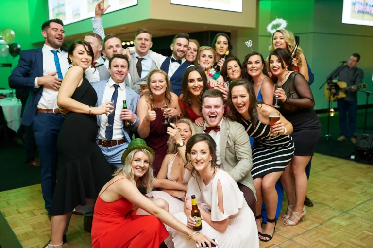 padraig pearses gac awards night mcg large group celebration party A large group of over 15 guests celebrate together during the Pearses Awards Night at the MCG, posing dynamically for the camera with laughter and cheers. Many hold beer bottles or wine glasses, while several women in elegant gowns crouch or lean in playfully. A central man laughs heartily with a drink in hand, and others raise hands in excitement. Behind them, a wooden dance floor, green ambient lighting, and a bar with live music create a lively, festive atmosphere, capturing the energy, camaraderie, and joyous revelry of the gala event.