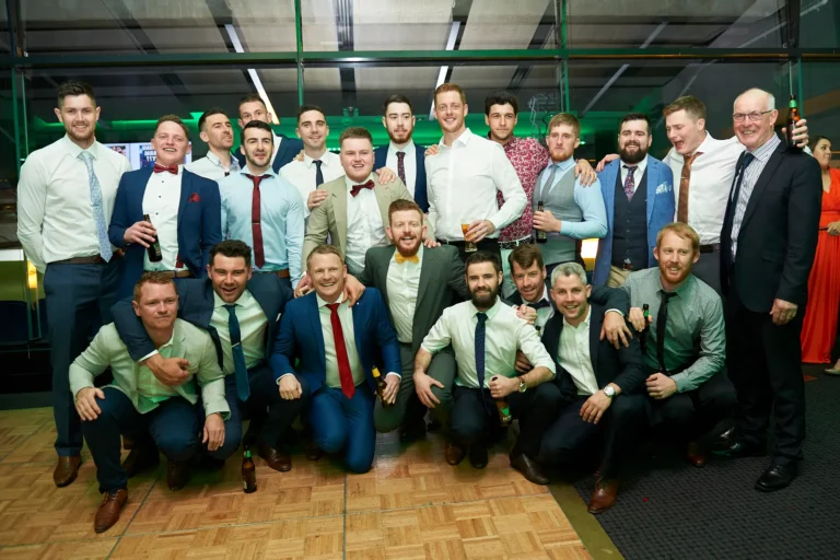 padraig pearses gac awards night mcg large group men posing A large group of over 15 men in formal attire pose together at the Pearses Awards Night at the MCG, with some standing and others crouching in the foreground. Many hold beer bottles while smiling warmly at the camera. The modern function room features glass partitions revealing stadium seating and the green MCG field beyond, with vibrant green ambient lighting reflecting off surfaces. Their relaxed, friendly postures and joyful expressions convey camaraderie, celebration, and the lively, festive atmosphere of the gala event.