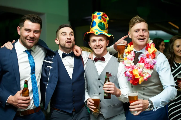 padraig pearses gac awards night mcg men party fun Four men in formal attire pose together during the Pearses Awards Night at the MCG, celebrating with broad smiles and playful accessories. The central man wears a colorful polka-dot party hat, while another sports a bright floral lei and raises a beer glass toward it. All hold drinks, enjoying the lively, festive moment. Behind them, a sleek modern event space with subtle green ambient lighting and glimpses of other guests adds to the energetic, social atmosphere of the gala party. The image captures camaraderie, fun, and joyful celebration among friends at the formal event.
