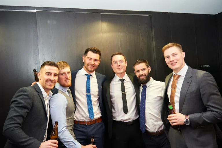 padraig pearses gac awards night mcg six men formal group smiling beers A group of six men dressed in formal suits stand closely together, smiling warmly at the camera during the Pearses Awards Night & Gala Dinner at the MCG. Several hold beer bottles, adding to the relaxed and social feel of the moment. Their friendly postures and shoulder-to-shoulder stance reflect genuine camaraderie as they enjoy the evening. Behind them, sleek dark wood-paneled walls and a small “ELECTRICAL” sign hint at a modern indoor area within the venue. The scene captures a lively, celebratory moment where friends pause to document their shared experience at the gala.