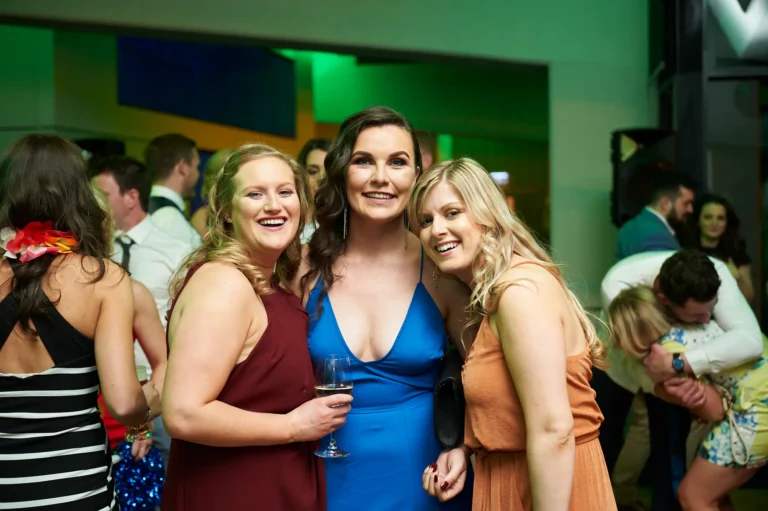 padraig pearses gac awards night mcg three women posing Three women pose together at the Pearses Awards Night at the MCG, smiling warmly at the camera. They wear elegant gowns in maroon, blue, and orange/peach, holding a wine glass and leaning in closely. The modern function room features dark interiors with green ambient lighting, while other guests mingle in the background. A candid moment of joy and lively social interaction highlights the festive and celebratory atmosphere of this gala event.