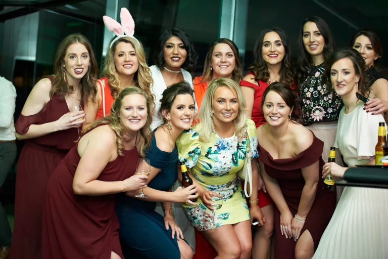 padraig pearses gac awards night mcg women celebration A group of ten women pose together during the Pearses Awards Night at the MCG, smiling warmly at the camera. Several hold wine glasses or SULME beer bottles, and one wears a pink bunny ear headband, adding a playful touch. Their elegant dresses and confident, relaxed postures convey friendship and joy. The indoor venue features dark walls with subtle modern lighting, creating a sophisticated, festive, and social atmosphere at the gala event.