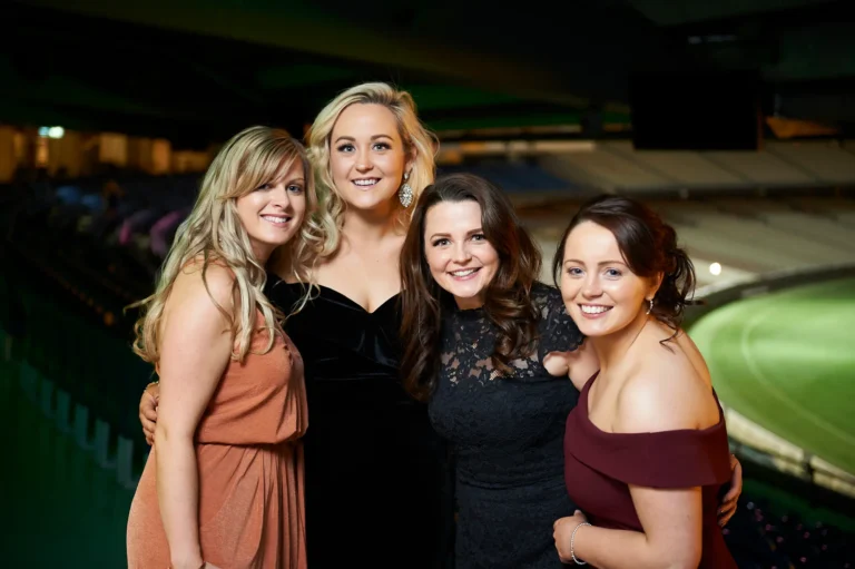 padraig pearses gac awards night mcg women group posing Four women pose together at the Pearses Awards Night at the MCG, smiling warmly at the camera with arms around each other’s waists or shoulders. They wear elegant gowns in peach, black velvet, black lace, and burgundy, conveying friendship and sophistication. Floor-to-ceiling glass partitions reveal dark blue stadium seating and the illuminated green MCG field, while green ambient lighting adds a festive and dynamic atmosphere to this celebratory gala moment.