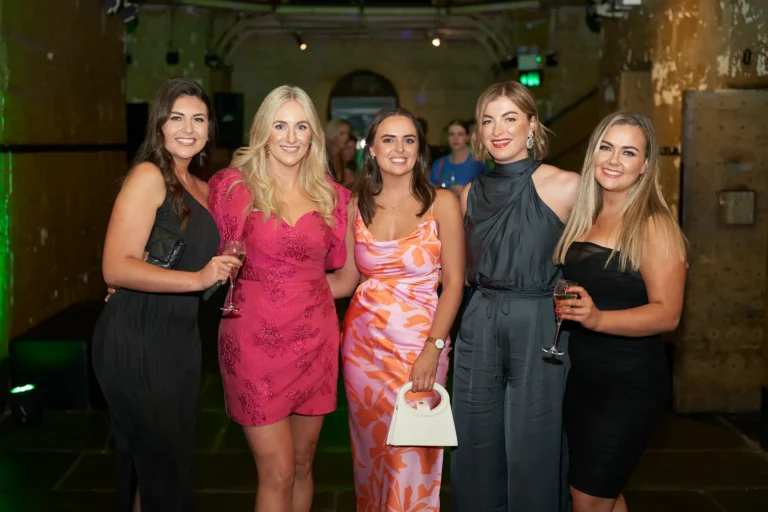 Five women pose together at the Padraig Pearses GAC Awards Night at Melbourne Old Gaol, smiling and holding wine glasses in a celebratory toast. The dimly lit historic bluestone walls with green uplighting create a sophisticated and lively atmosphere, while elegantly dressed guests mingle in the background, capturing the social and festive energy of the evening.