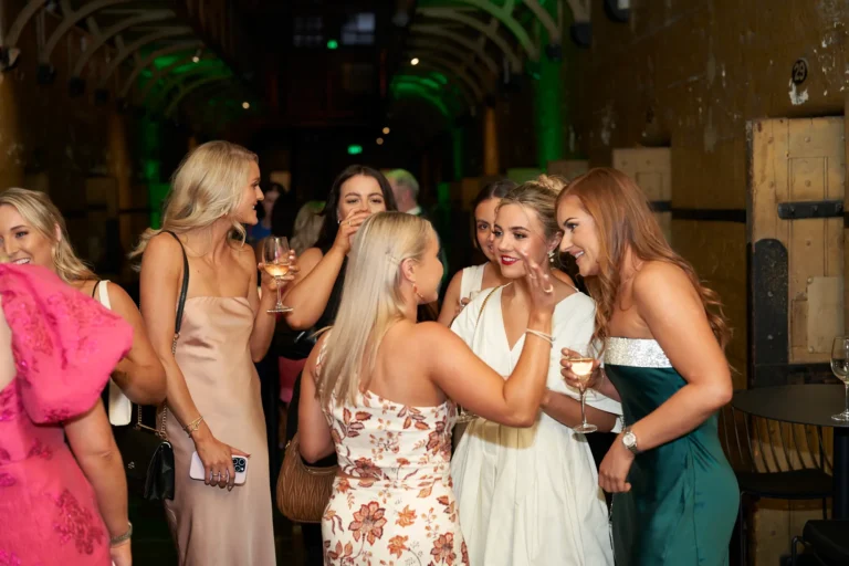 Five women share a joyful moment at the Padraig Pearses GAC Awards Night at Melbourne Old Gaol, holding wine glasses and smiling warmly as they interact and celebrate together. Set against the rugged yellow-toned bluestone walls of the historic venue with green uplighting, the image captures the sophisticated yet relaxed energy of the evening, where elegantly dressed guests mingle, laugh, and enjoy drinks, highlighting the festive and memorable atmosphere of this community-focused year-end celebration.