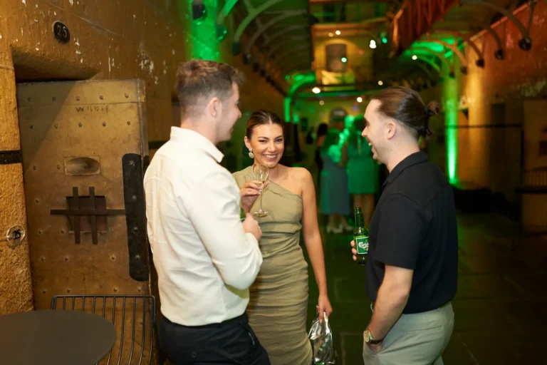 Two men and one woman engage in lively conversation at the Padraig Pearses GAC Awards Night at Melbourne Old Gaol, holding drinks and smiling naturally. Set against the rugged bluestone walls and a large metal cell door, with dramatic green uplighting highlighting the arched hallway, the image captures the sophisticated yet relaxed energy of the evening. Elegantly dressed guests mingle in the background, while the historic venue’s architectural details create a memorable and immersive atmosphere, blending formal celebration with the unique charm of this iconic Melbourne location.
