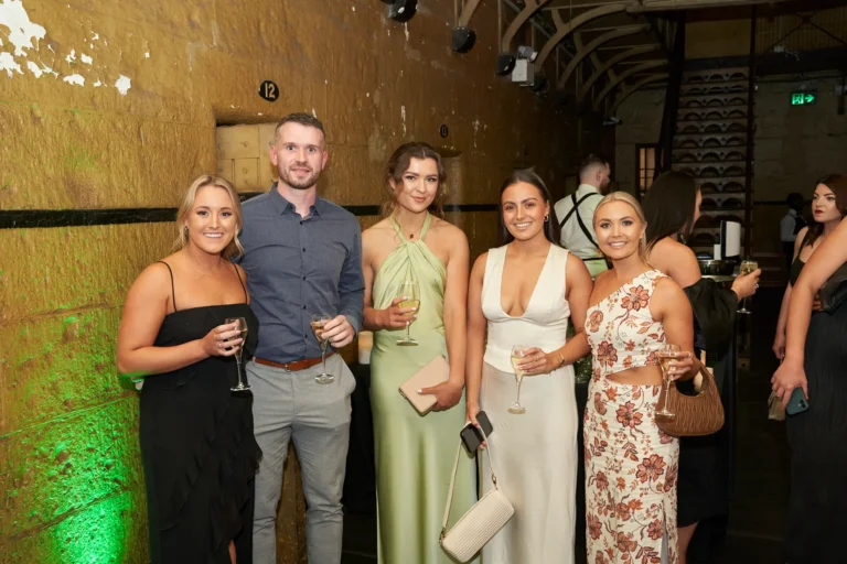 Five guests pose together at the Padraig Pearses GAC Awards Night at Melbourne Old Gaol, smiling warmly and holding wine glasses in a celebratory toast. The dimly lit historic bluestone walls and green uplighting create a sophisticated, lively atmosphere, while other elegantly dressed attendees mingle in the background, highlighting the festive and social energy of the event.