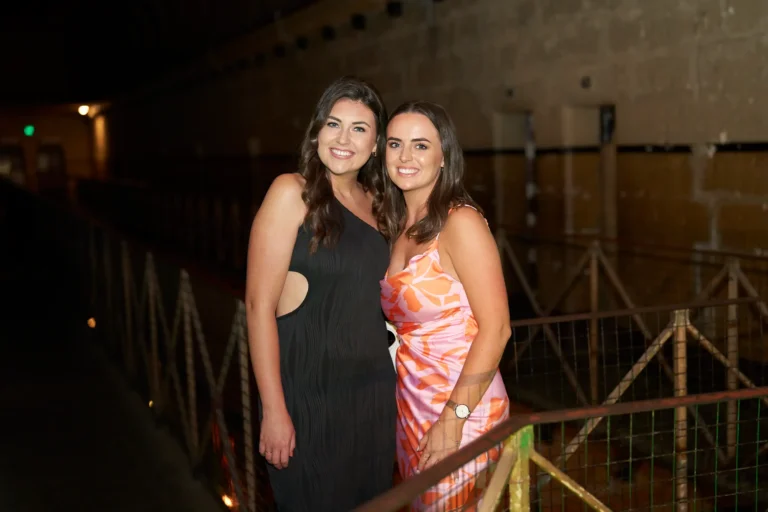 Two women pose together at the Padraig Pearses GAC Awards Night at Melbourne Old Gaol, smiling warmly while enjoying the evening. Standing on a metal walkway against rugged stone walls and industrial railings, the dimly lit venue is accented with strategic event lighting that highlights its historic architecture. Elegantly dressed in a black cutout midi dress and a pink-orange patterned slip dress, the women reflect the sophisticated, social, and celebratory energy of the awards night, while the unique historic setting creates a memorable backdrop for candid interactions and festive moments.