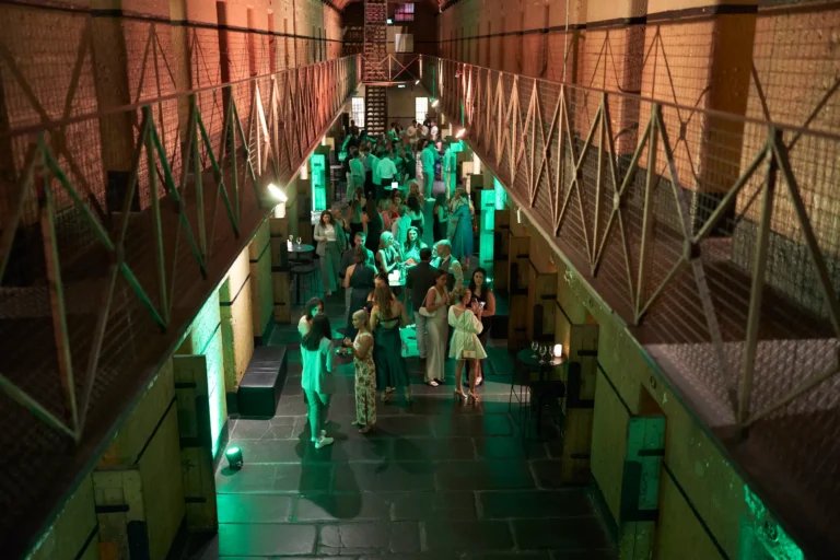 A lively crowd of 30–40 elegantly dressed guests mingle and socialize at the Padraig Pearses GAC Awards Night held at Melbourne Old Gaol. Groups hold wine glasses and beer bottles while chatting casually across the dimly lit main cell block. Rugged bluestone walls, exposed metal catwalks, and dramatic green uplighting highlight the historic architecture, creating a sophisticated, festive, and energetic atmosphere. The combination of relaxed interactions, flowing drinks, and the unique moody setting of this former prison forms a memorable backdrop for the evening’s celebratory awards night and end-of-year gathering.