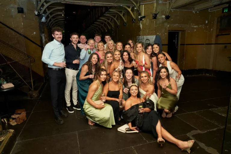 A large group of around 25 guests pose together during the Pearses Awards Night & End of Year Celebration at Melbourne Old Gaol. Men and women stand and crouch in relaxed rows, smiling and holding drinks as they enjoy the moment. Rugged bluestone walls, exposed metal trusses, a visible catwalk, and subtle green uplighting create a dramatic historic backdrop. The image captures the energy, camaraderie, and shared celebration of the evening within the iconic former prison venue.