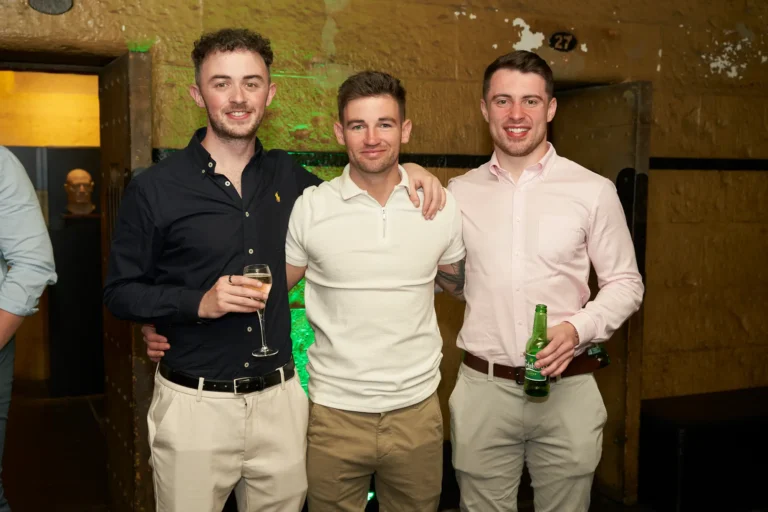 Three men pose together at the Padraig Pearses GAC Awards Night at Melbourne Old Gaol, smiling warmly with the central man’s arms around his friends’ shoulders, reflecting camaraderie and celebration. One holds a champagne flute, another a green beer bottle, while the rugged yellow-toned bluestone walls, numbered metal doors, and vibrant green uplighting create a moody, historic backdrop. Elegantly dressed guests mingle in the background, highlighting the lively, sophisticated, and festive atmosphere. The combination of relaxed interactions, formal attire, and the iconic former prison setting captures the memorable and distinctive energy of this end-of-year awards night.