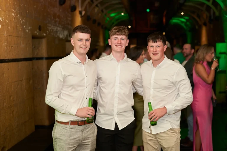 Three young men stand side by side at the Padraig Pearses GAC Awards Night at Melbourne Old Gaol, smiling warmly and holding green beer bottles in a relaxed, celebratory pose. Dressed in white button-down shirts and formal trousers, they are framed by rugged yellow-toned bluestone walls and arched ceilings highlighted with green uplighting. Elegantly dressed guests mingle on the catwalk above, creating a lively and social scene. The historic architecture of the former prison, combined with the festive energy and sophisticated attire, provides a distinctive, moody, and memorable backdrop for this end-of-year awards night celebration.