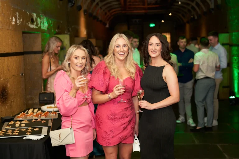 Three women pose together at the Padraig Pearses GAC Awards Night at Melbourne Old Gaol, smiling warmly and holding wine glasses in a celebratory toast. Standing near a buffet table with food platters, they are set against rugged yellow-toned bluestone walls highlighted by vibrant green uplighting in the arches. Elegantly dressed guests mingle in the dimly lit historic venue, creating a lively, sophisticated, and festive atmosphere. The image captures the social energy and joyful interactions of the evening, with the unique architecture and moody historic setting providing a memorable and distinctive backdrop for the awards night celebration.