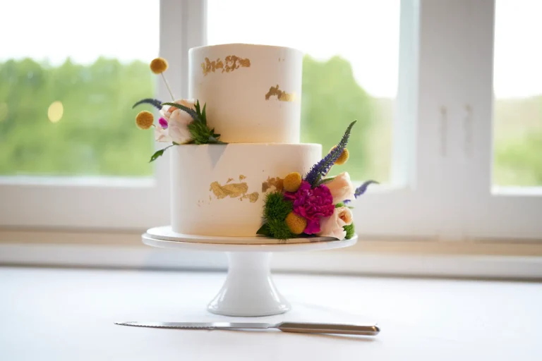 A two-tiered white wedding cake with gold leaf accents and fresh flowers, including pink peonies, magenta carnations, yellow craspedia, and purple spiky blooms, sits elegantly on a white cake stand at Farm Vigano. A stainless steel cake knife rests in the foreground. Large white-framed windows reveal a blurred green garden and Plenty Gorge Valley, with natural light streaming in. The scene highlights a sophisticated yet rustic cake display, blending modern elegance with the venue’s lush, Italian-inspired surroundings, ready for the couple and guests to celebrate this traditional wedding moment.