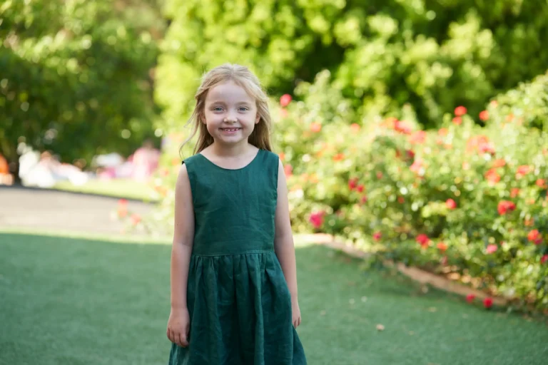 A young flower girl in a dark green sleeveless dress smiles brightly at the camera during Allison and Christian’s outdoor wedding at Farm Vigano in South Morang, Melbourne. She stands on a manicured lawn surrounded by lush greenery, mature trees, and a vibrant red-flowered bush, with natural sunlight enhancing the cheerful, innocent, and relaxed atmosphere. The scene captures a sweet, candid moment, highlighting the charm and elegance of the venue’s picturesque gardens.