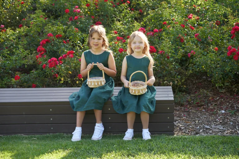 Two young flower girls in matching dark green dresses and white sneakers sit side-by-side on a low bench at Allison and Christian’s pre-ceremony moments at Farm Vigano in South Morang, Melbourne. Each holds a small wicker basket, likely filled with petals or confetti, framed by a lush bush of vibrant red roses and greenery. Bathed in bright natural daylight on the manicured lawn, the scene captures innocence, cheerfulness, and a relaxed, charming atmosphere in the picturesque outdoor setting.