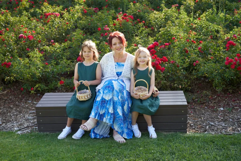 A woman guest in a blue and white patterned dress with a white cardigan sits on a dark brown bench between two young flower girls in matching dark green dresses and white sneakers during pre-ceremony moments at Allison and Christian’s wedding at Farm Vigano in South Morang, Melbourne. Each girl holds a small wicker basket of petals or confetti, framed by vibrant red roses and lush greenery. Bathed in bright natural sunlight on the manicured lawn, the scene captures a cheerful, relaxed, and intimate atmosphere full of warmth and anticipation.