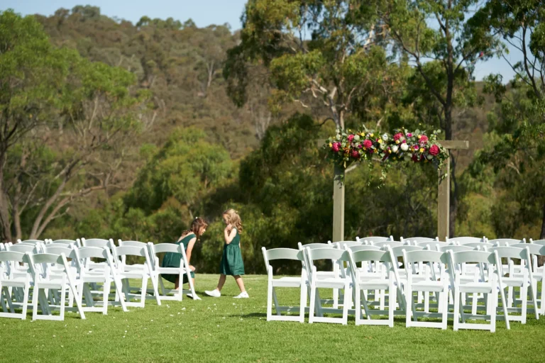 Two young flower girls in dark green dresses share a candid moment on the manicured lawn at Farm Vigano in South Morang, Melbourne, moments before the outdoor wedding ceremony. They are surrounded by neatly arranged white chairs and a rustic wooden arch adorned with red, white, and yellow flowers, with dense greenery and distant hills in the background. The sun-drenched, tranquil scene captures innocence, anticipation, and natural elegance, highlighting a charming and intimate pre-ceremony moment in a picturesque setting.
