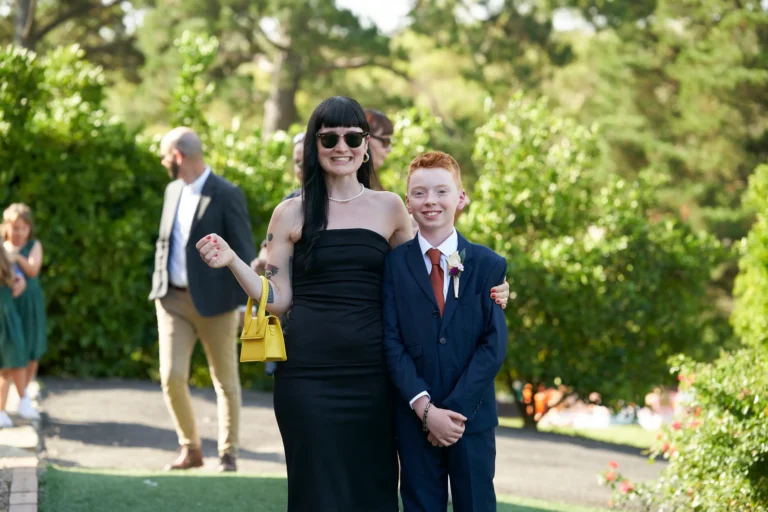 A joyful, sunlit moment at Allison and Christian’s outdoor wedding at Farm Vigano in South Morang, Melbourne, showing a woman in a black strapless dress with a yellow handbag smiling at the camera alongside a young boy in a navy suit and rust-colored tie. A man and another child are captured in candid motion nearby, set against lush gardens and mature trees, creating a relaxed, elegant, and cheerful atmosphere in the bright natural light, reflecting the warm and personal celebration of the day.