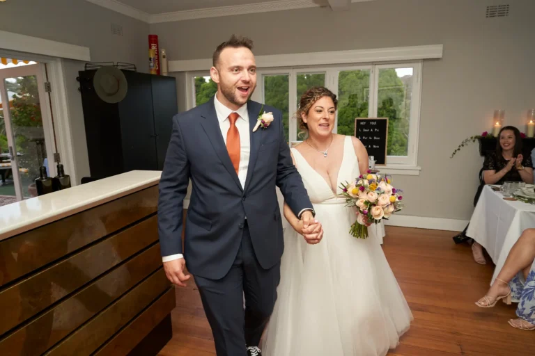 The bride and groom make their joyful entrance at their wedding reception at Farm Vigano, walking hand-in-hand across polished hardwood floors. The groom wears a navy suit with a rust-colored tie, and the bride wears a white V-neck gown holding a multi-colored bouquet. Guests seated at white-linened tables clap and smile warmly in the background. A dark wood and marble bar is visible to the left, while large white-framed windows showcase lush green gardens outside. The scene is bright, lively, and welcoming, capturing the excitement and celebration of the couple's grand reception arrival.