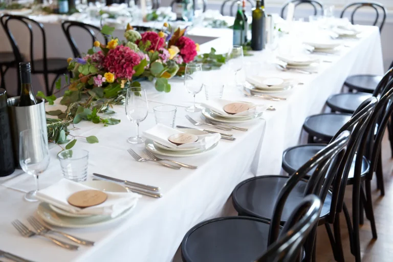 An elegant and rustic wedding reception table at Farm Vigano, featuring crisp white linen, dark bentwood chairs, and a flowing greenery garland adorned with vibrant magenta hydrangeas, yellow blooms, and eucalyptus. Each place setting includes white plates, glassware, stainless cutlery, and a natural wood place card. Wine bottles and a black metallic cooler are arranged along the table. The indoor space with wooden floors and other tables in the background combines rustic charm with sophisticated Italian-inspired details, creating a warm, inviting, and celebratory atmosphere for guests.