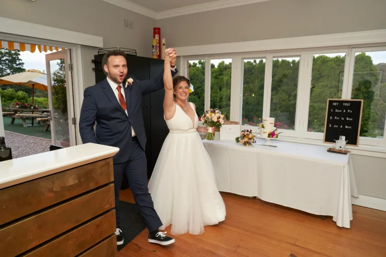 The bride and groom make a lively, triumphant entrance at their wedding reception at Farm Vigano, walking hand-in-hand with their arms raised in excitement. The groom wears a navy suit, rust-colored tie, and black sneakers, while the bride wears a white V-neck gown holding a multi-colored bouquet. Guests look on and smile as they enter the indoor space with polished hardwood floors, white-framed windows overlooking lush gardens, and a dark wood marble-topped bar. A display table with a two-tiered wedding cake, wicker basket, and interactive guest sign adds charm. The scene is energetic, warm, and celebratory.