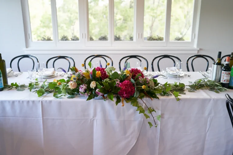 An elegant and rustic wedding reception table setup at Farm Vigano, featuring a long table covered with crisp white linens and surrounded by black bentwood-style chairs. A lush greenery garland runs along the table, with natural light streaming through large white-framed windows overlooking a vibrant garden with mature trees. The bright, airy indoor space blends rustic charm with sophistication, creating a warm and inviting atmosphere for guests, highlighting the Italian-inspired elegance and the venue’s beautiful natural surroundings.