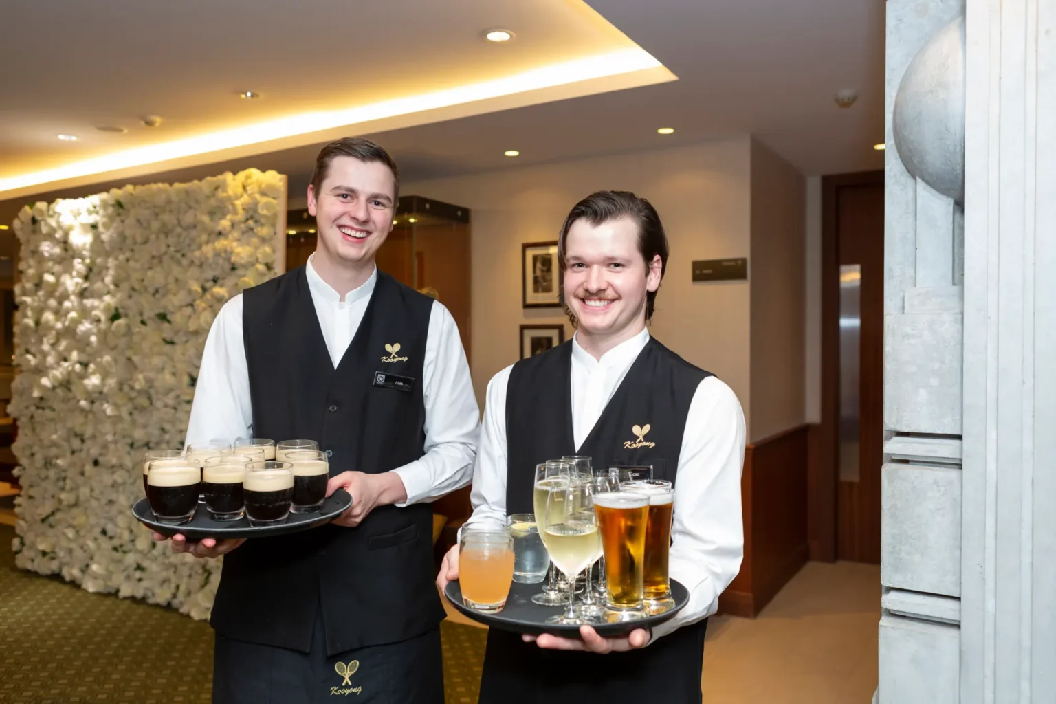 Two male food and beverage attendants pose at the Kooyong Lawn Tennis Club End-of-Year Masquerade Ball in front of a white and cream rose floral wall. Dressed in white shirts, black vests with club logos, and dark trousers, each holding a tray of drinks including champagne, beer, and cocktails, they smile confidently at the camera, reflecting the professional, sophisticated, and celebratory atmosphere of this prestigious Melbourne end-of-year event.