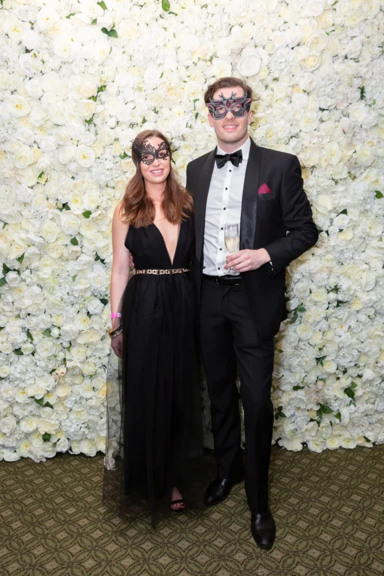 kooyong lawn tennis club end of year masquerade ball elegant arrivals couple black A couple poses together at the Kooyong Lawn Tennis Club End-of-Year Masquerade Ball in front of a white and cream rose floral wall. Dressed in elegant black formalwear, the man in a classic tuxedo with a black bow tie and the woman in a deep V-neck black gown with a chain-link belt, both wearing masquerade eye masks, they smile confidently at the camera, capturing the glamorous, sophisticated, and celebratory atmosphere of this prestigious Melbourne end-of-year event.
