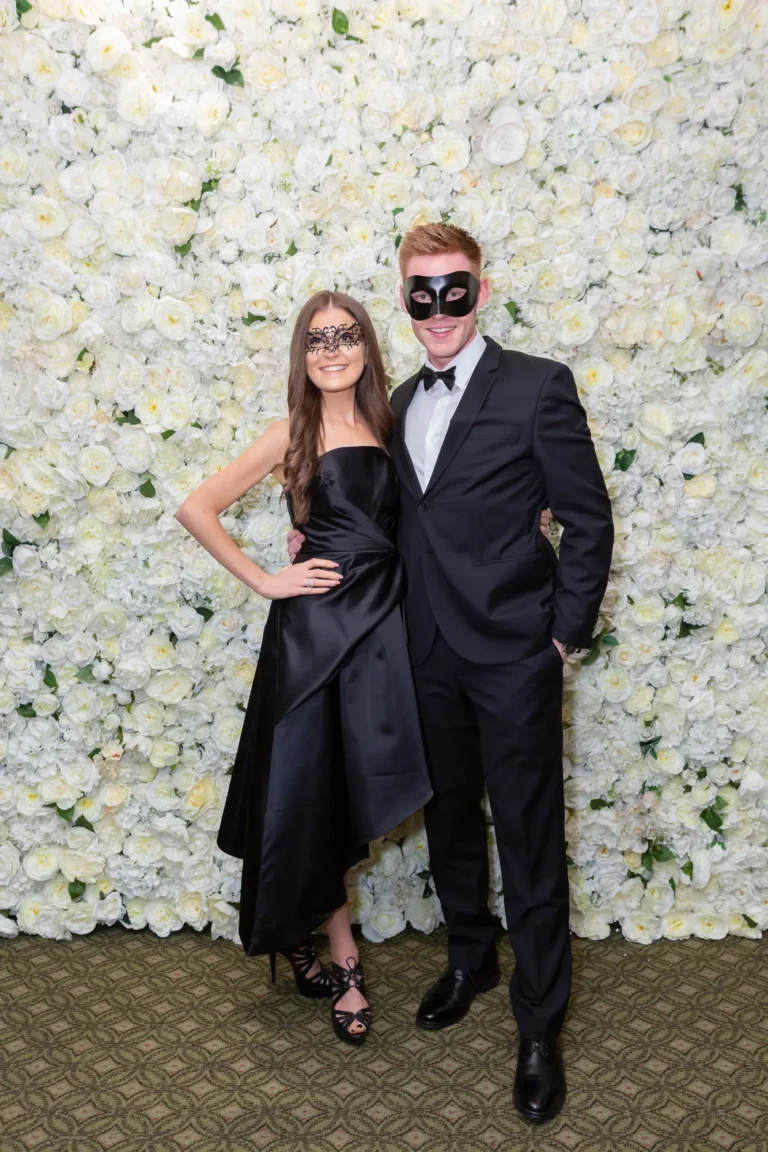 kooyong lawn tennis club end of year masquerade ball elegant arrivals couple black dress A couple poses together at the Kooyong Lawn Tennis Club End-of-Year Masquerade Ball in front of a white and cream rose floral wall. Dressed in elegant black formalwear, the man in a tuxedo with a black bow tie and masquerade eye mask and the woman in a black strapless gown with an asymmetric hem and lace eye mask, they smile confidently at the camera, capturing the sophisticated, festive, and glamorous atmosphere of this prestigious Melbourne end-of-year masquerade event.