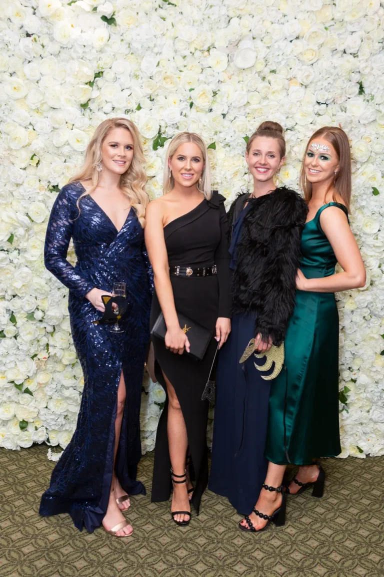 kooyong lawn tennis club end of year masquerade ball elegant arrivals four women Four women pose together at the Kooyong Lawn Tennis Club End-of-Year Masquerade Ball in front of a white and cream rose floral wall. Dressed in formal evening gowns, sequins, stylish cocktail dresses, and masquerade accessories including masks and face paint, they smile confidently at the camera, capturing the social sophistication, glamour, and celebratory atmosphere of this prestigious Melbourne end-of-year masquerade event.