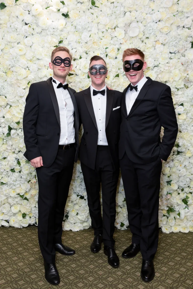 kooyong lawn tennis club end of year masquerade ball elegant arrivals three men Three men pose together on arrival at the Kooyong Lawn Tennis Club End-of-Year Masquerade Ball, standing in front of a white and cream rose floral wall. Dressed in classic black-tie tuxedos and masquerade eye masks, they smile confidently at the camera, capturing the refined social energy, camaraderie, and sense of occasion that defines this prestigious Melbourne end-of-year celebration.