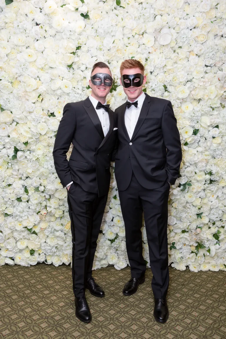 kooyong lawn tennis club end of year masquerade ball elegant arrivals two men Two men arrive and pose together at the Kooyong Lawn Tennis Club End-of-Year Masquerade Ball, standing in front of a white and cream rose floral wall. Dressed in classic black-tie tuxedos with bow ties and masquerade eye masks, they smile confidently at the camera, reflecting the refined, social atmosphere and sense of anticipation that defines guest arrivals at this prestigious Melbourne end-of-year celebration.