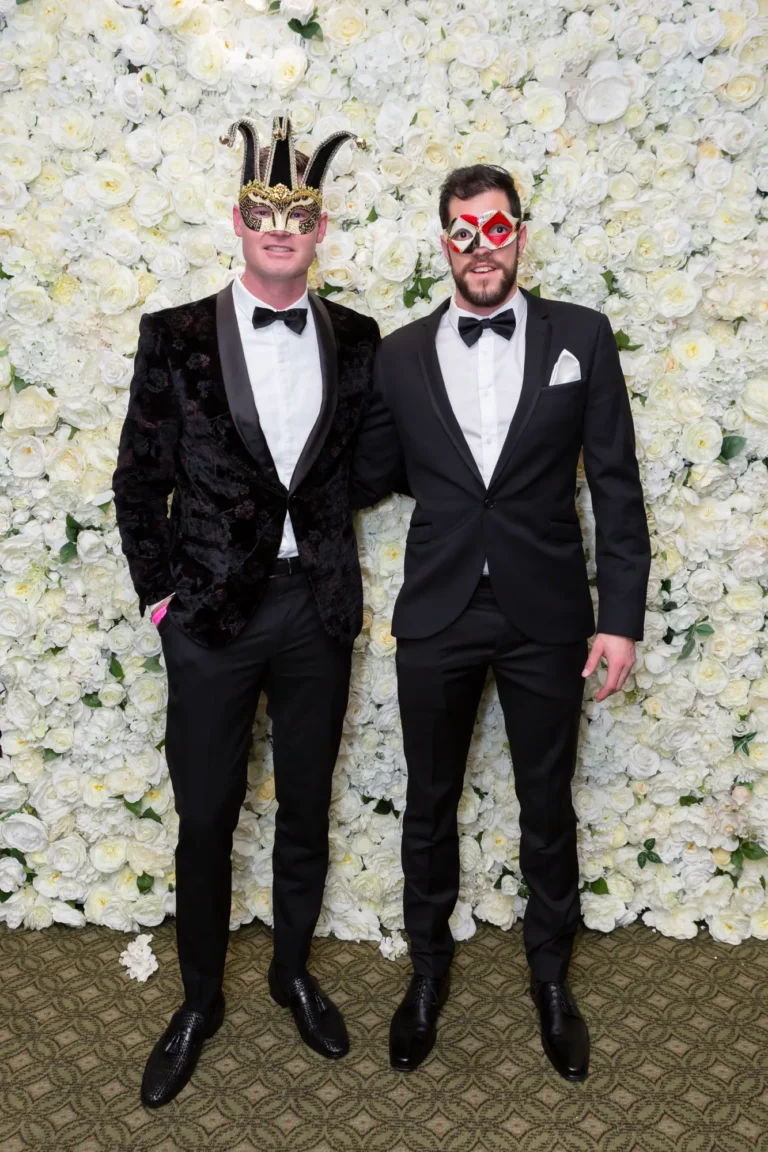 kooyong lawn tennis club end of year masquerade ball elegant arrivals two men masks Two men pose together at the Kooyong Lawn Tennis Club End-of-Year Masquerade Ball in front of a white and cream rose floral wall. Dressed in elegant black tuxedos with masquerade eye masks—one in a velvet double-breasted jacket and the other in a single-button tuxedo with a Harlequin-style mask—they smile confidently at the camera, reflecting the sophisticated, celebratory, and glamorous atmosphere of this prestigious Melbourne end-of-year masquerade event.