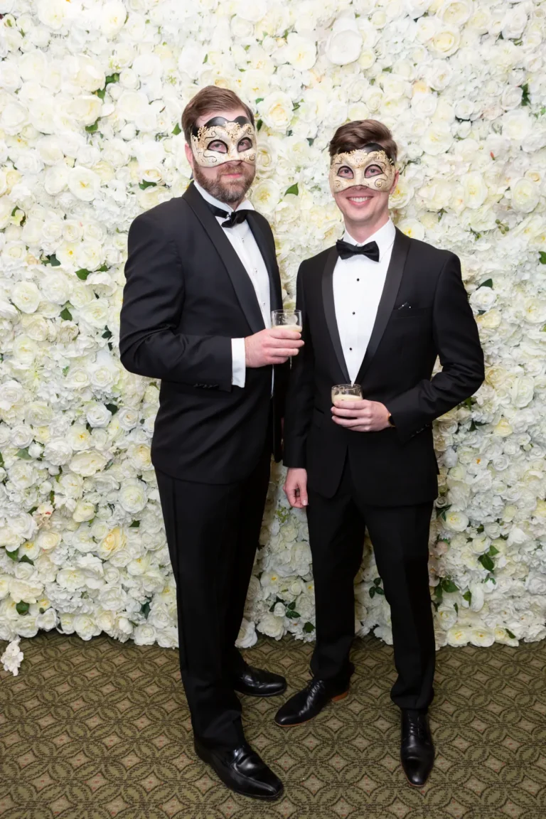 kooyong lawn tennis club end of year masquerade ball elegant arrivals two men tuxedos Two men pose together at the Kooyong Lawn Tennis Club End-of-Year Masquerade Ball in front of a white and cream rose floral wall. Dressed in black tuxedos with bow ties and masquerade eye masks, each holding a glass, they smile confidently at the camera, capturing the sophisticated, celebratory, and glamorous atmosphere of this prestigious Melbourne end-of-year masquerade event.
