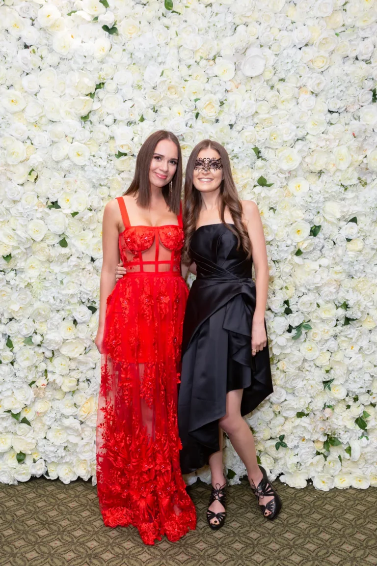 kooyong lawn tennis club end of year masquerade ball elegant arrivals two women red black gowns Two women arrive and pose together at the Kooyong Lawn Tennis Club End-of-Year Masquerade Ball, standing before a white and cream rose floral wall. Dressed in elegant formal gowns, one in a flowing red lace dress and the other in a black strapless gown with a lace eye mask, they smile warmly at the camera, reflecting the refined glamour, social energy, and celebratory spirit of this prestigious Melbourne end-of-year masquerade event.