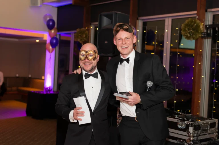 kooyong lawn tennis club end of year masquerade ball hosts speeches A prize winner stands beside the event host during speeches at the Kooyong Lawn Tennis Club End-of-Year Masquerade Ball in Melbourne, both dressed in black-tie tuxedos and masquerade masks, posing near the DJ and sound equipment under soft purple lighting, capturing a celebratory moment of recognition during the elegant end-of-year event.