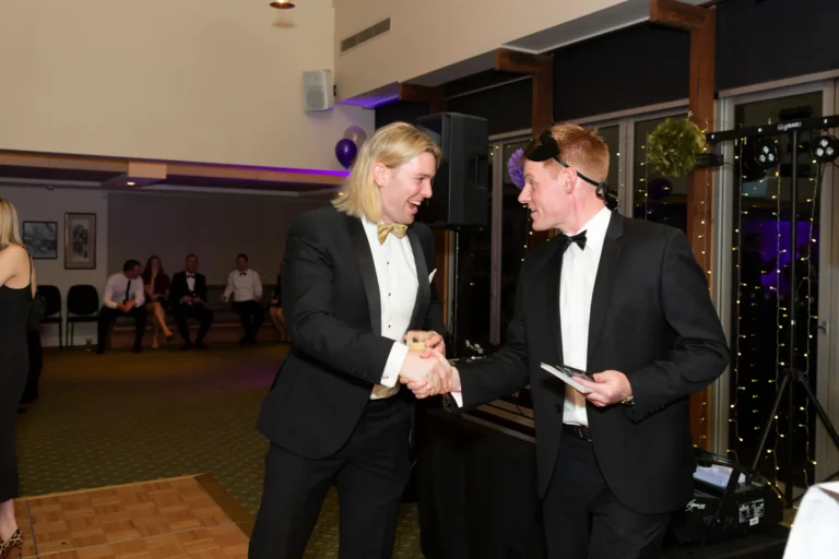 kooyong lawn tennis club masquerade ball award presentation A prize winner shakes hands with the event speaker during an awards presentation at the Kooyong Lawn Tennis Club End-of-Year Masquerade Ball in Melbourne. Both men are dressed in black-tie tuxedos with masquerade eye masks, smiling warmly as the winner holds a small trophy. The indoor venue features green-patterned carpet, polished wooden floors, purple and gold balloon clusters, framed artwork, and professional audio equipment, capturing a festive, elegant, and celebratory moment during the speeches and formal proceedings.