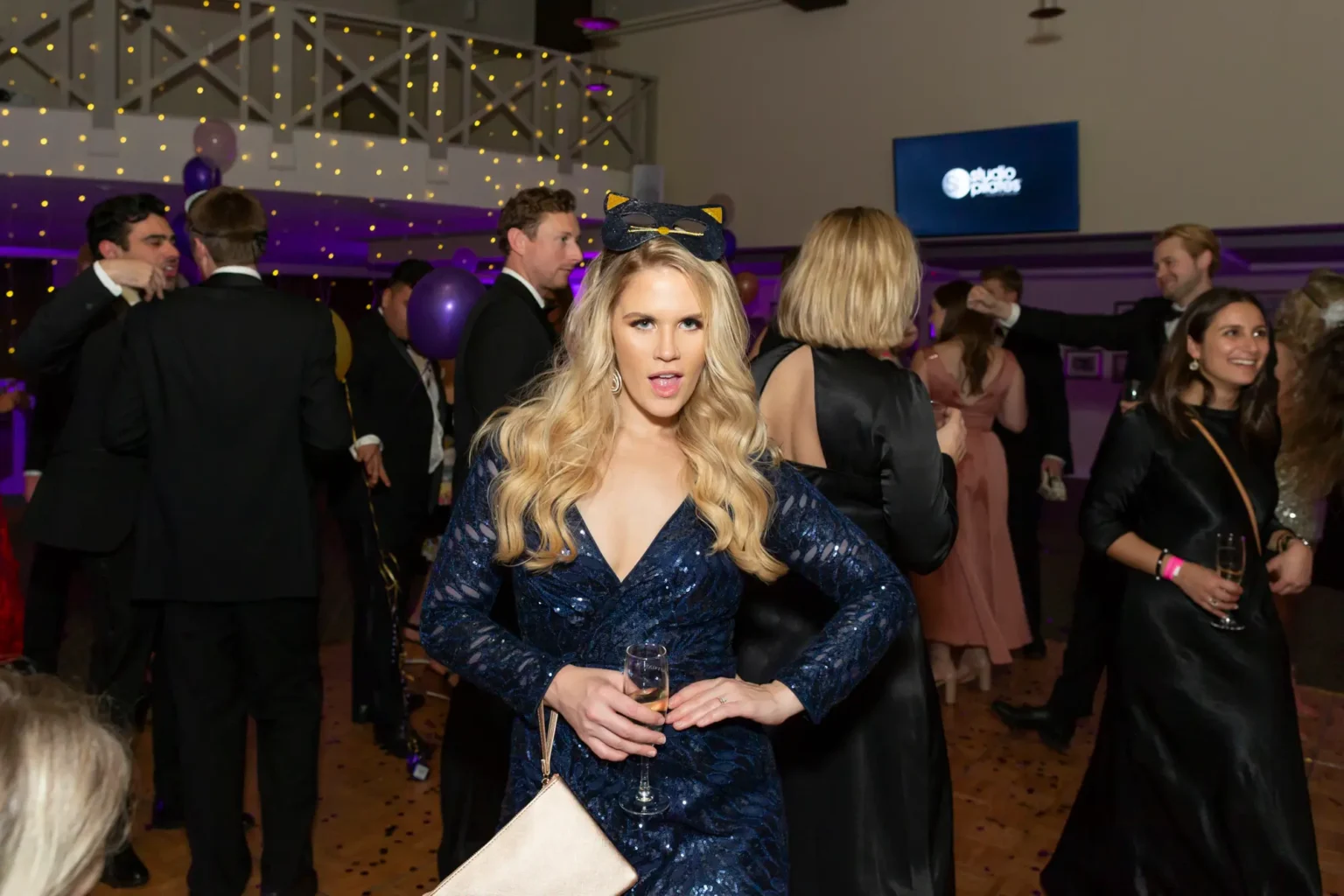 A woman in a deep blue sequined dress strikes a playful pose wearing a black cat-ear mask at the Kooyong Lawn Tennis Club End-of-Year Masquerade Ball. Holding a champagne flute and clutch, she smiles energetically toward the camera as guests in tuxedos and cocktail dresses mingle behind her beneath fairy lights and purple balloons, capturing the glamorous and lively atmosphere of the celebration.