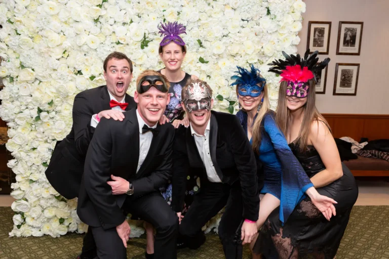 kooyong lawn tennis club masquerade ball candid group flower wall celebration Six guests pose playfully in front of a white flower wall at the Kooyong Lawn Tennis Club Masquerade Ball in Toorak, wearing tuxedos, elegant dresses, and ornate masks. Their relaxed, joyful expressions capture the lively, glamorous spirit of the YarraYong Ball inside the club’s elegant function room.