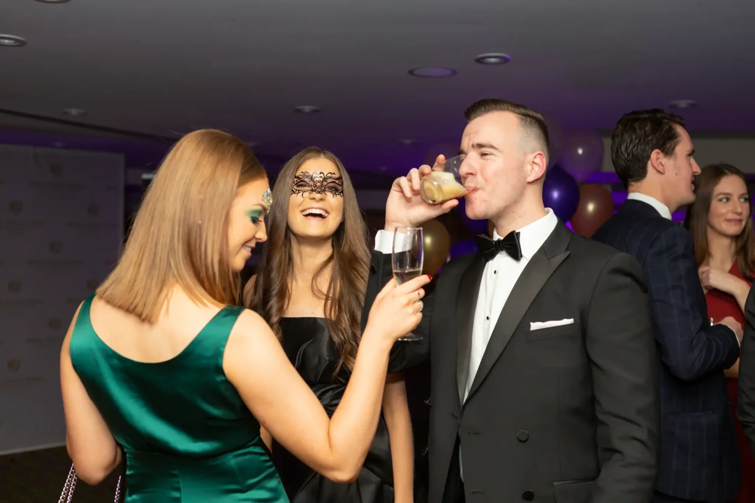 Three guests are captured in a lively, candid moment at the Kooyong Lawn Tennis Club End-of-Year Masquerade Ball in Toorak. A man in a tuxedo drinks from a pale yellow cocktail, while a woman in a black dress with an intricate black lace mask laughs joyfully beside him. A woman in a green satin dress, seen from behind, holds a champagne flute, engaging with the group. In the mid-ground, a man in a blue textured suit and a woman in a red dress converse. The indoor venue features ambient purple lighting, clusters of balloons, and other formally dressed guests, creating a sophisticated, festive, and celebratory atmosphere.