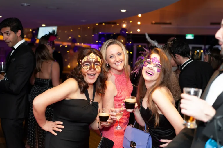 kooyong lawn tennis club masquerade ball candid group with purple foil and glass partition A joyful group captured at the Kooyong Lawn Tennis Club YarraYong Ball in Toorak, featuring three women in elegant black and coral evening dresses with intricate masquerade masks, smiling for the camera. Two men in black tuxedos are partially visible on either side, one holding a glass of beer. The backdrop includes shimmering purple and silver foil curtains, a glass partition, and ambient purple lighting, highlighting the festive and sophisticated atmosphere of the indoor end-of-year celebration.
