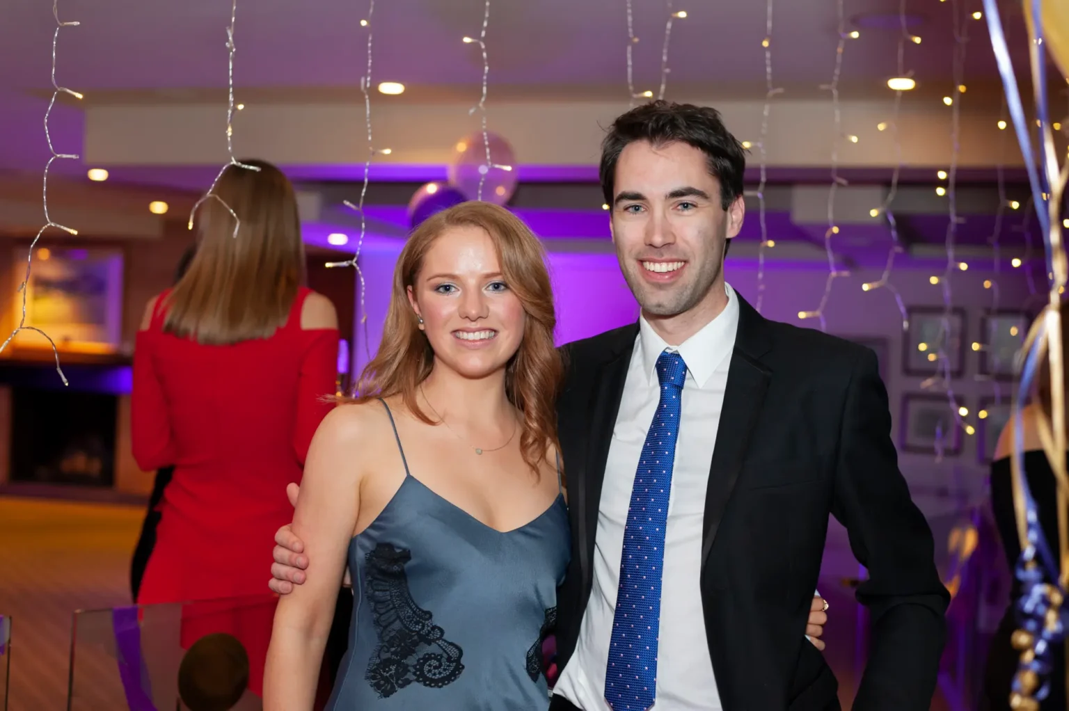 A man and woman pose arm-in-arm at the YarraYong Ball at Kooyong Lawn Tennis Club in Toorak, smiling broadly and enjoying the festive evening. The woman wears a grey-blue dress with lace detailing, and the man wears a black suit with a bright blue tie. Warm ambient lighting, string lights, and balloons create a celebratory and sophisticated end-of-year ball atmosphere.