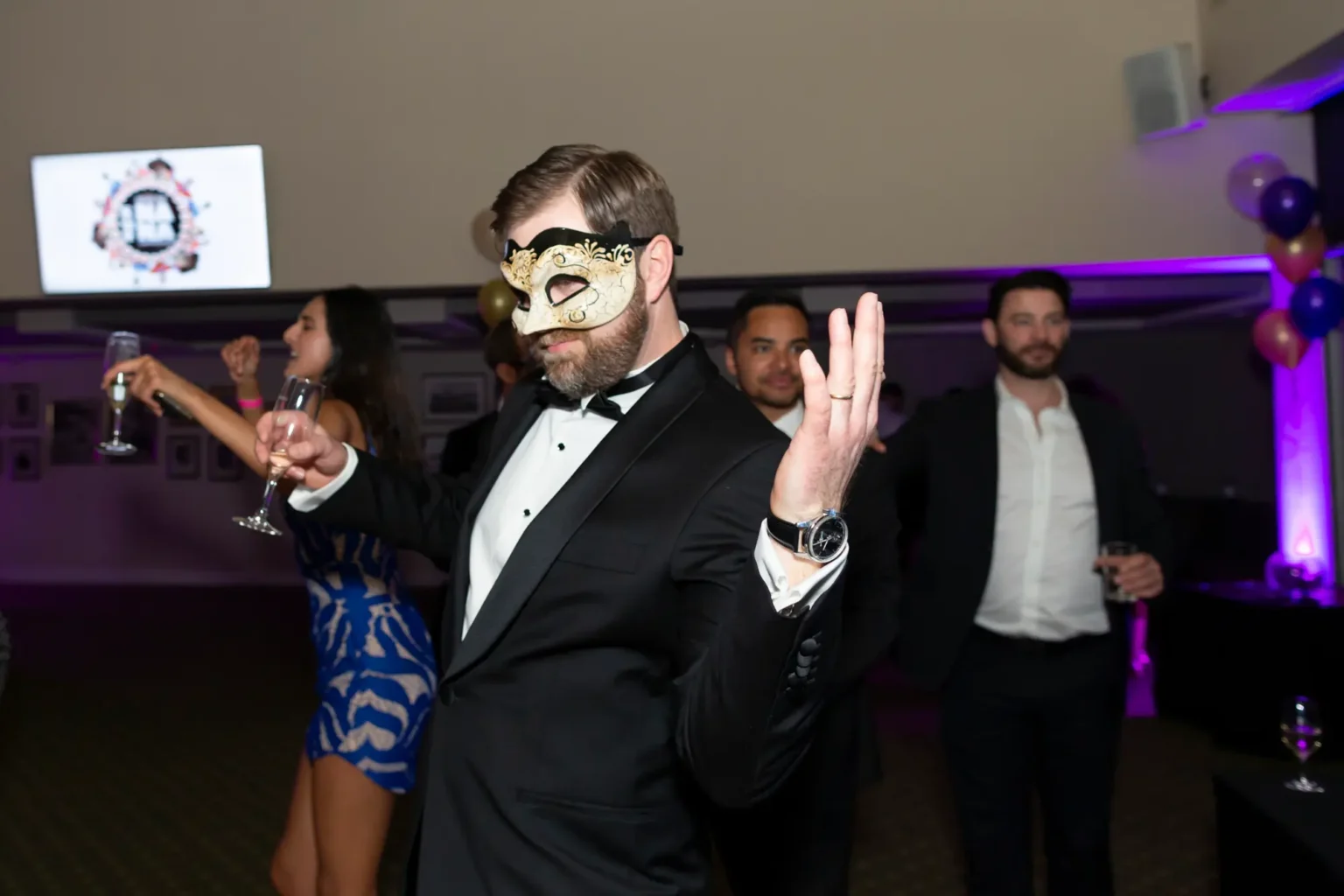 Three guests enjoy lively moments at the Kooyong Lawn Tennis Club End-of-Year Masquerade Ball in Toorak. A man in the foreground wearing a black and gold masquerade mask gestures enthusiastically while holding a champagne glass, accompanied by another smiling man in a tuxedo and a woman in a blue-and-white patterned dress raising her glass. The indoor venue is decorated with purple and gold balloons, string lights, and ambient purple lighting, creating a festive and celebratory atmosphere.